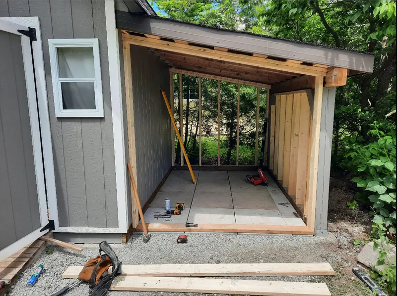 An open-air covered extension is framed onto the side of a shed, with a concrete pad below.