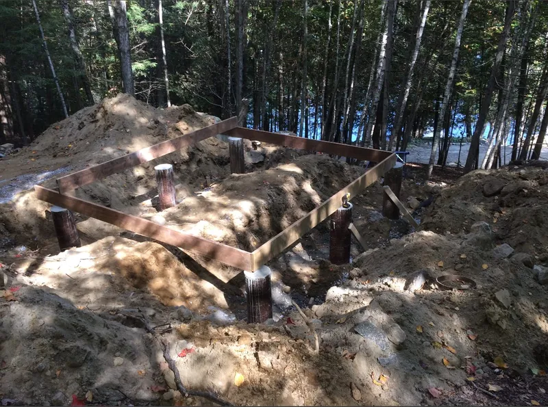 A floor frame rests on pier footings, with trees and water visible beyond.