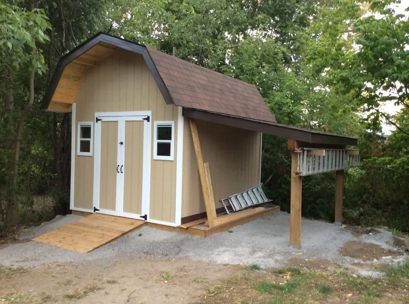 A finished tan shed with a gambrel roof, a ramp, and an attached covered deck with railing.